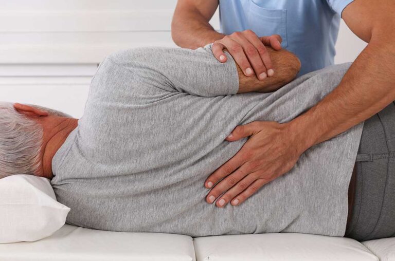 Person gently stretching their lower back beside a chair in a bright home.